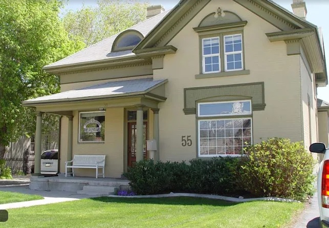 A beige two-story house at Parkside Apartments Provo with a covered front porch, white bench, green trim, large windows, and the number 55 on the exterior, surrounded by grass, bushes, and trees.