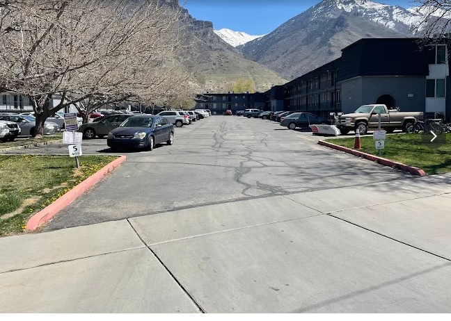 A parking lot with several parked cars at Heritage Court Apartments Provo, bordered by leafless trees and buildings. Snow-capped mountains are visible in the background under a clear sky.