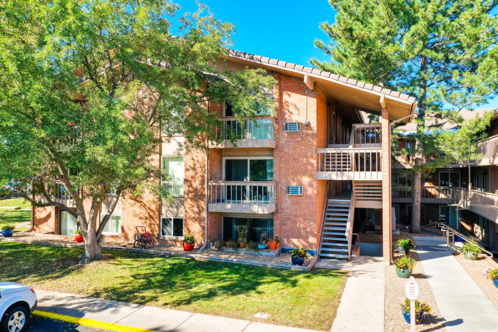 A three-story brick apartment building, Coronado Apartments Boulder, features balconies, outdoor stairs, mature trees, potted plants, and a bright blue sky above. A white car is parked in front of the building.