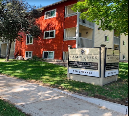 A red and beige apartment building with a sign in front reading Campus Park Apartments Fort Collins and listing contact information, surrounded by grass, trees, and a sidewalk on a sunny day.