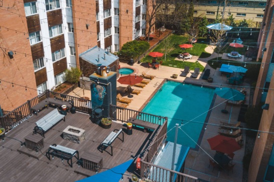Aerial view of Sterling Boulder Apartments Boulder featuring a rooftop lounge area, swimming pool with lounge chairs, hot tub, and patio tables with umbrellas, all surrounded by brick buildings and green lawns.