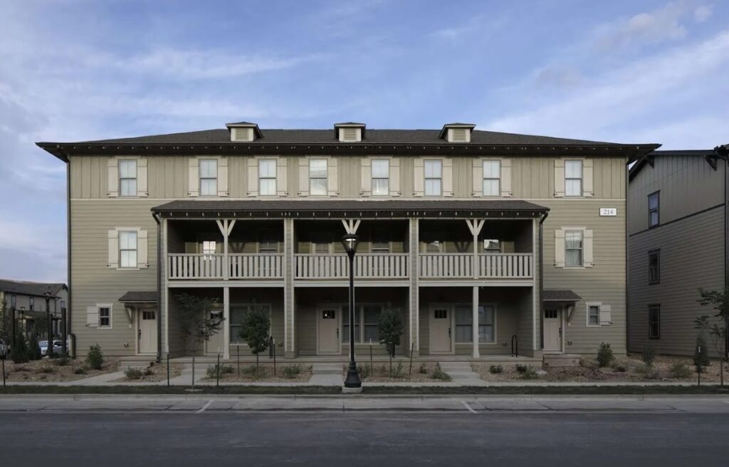 A two-story beige apartment building at The Cottages of Fort Collins with white shutters and a covered front porch, featuring four entry doors, balconies, a lamppost in front, and a landscaped area with small trees and shrubs.