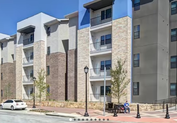 A modern apartment building, Campus Edge on UTA Boulevard Arlington, features brick and light-colored facades, balconies, and a person relaxing on a bench near the sidewalk and parked white car under a clear blue sky.