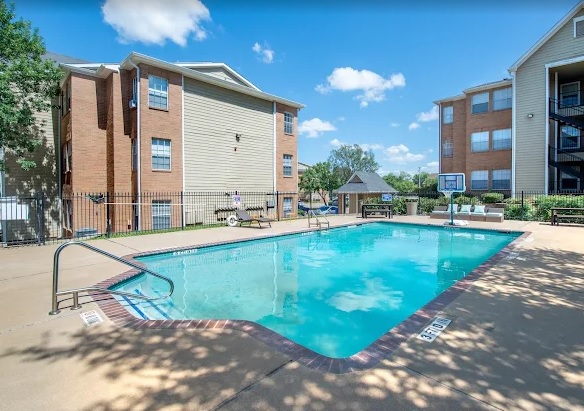 Outdoor swimming pool surrounded by a concrete deck, lounge chairs, and benches at Centennial Court Apartments Arlington, with apartment buildings and a gazebo in the background under a sunny, blue sky with scattered clouds.