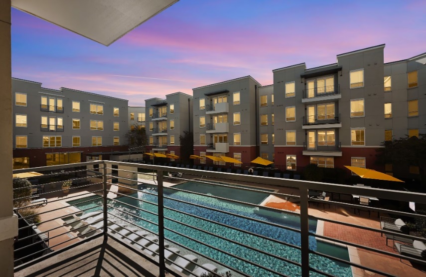 Enjoy the view from your Liv+ Arlington balcony, overlooking a pool and lounge area framed by modern apartments at dusk, glowing windows, and a vibrant sunset sky in the background.