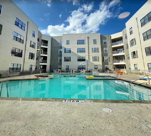 A large outdoor swimming pool at Maverick Place Apartments Arlington is surrounded by a multi-story building with balconies, lounge chairs, and a blue sky with scattered clouds above.