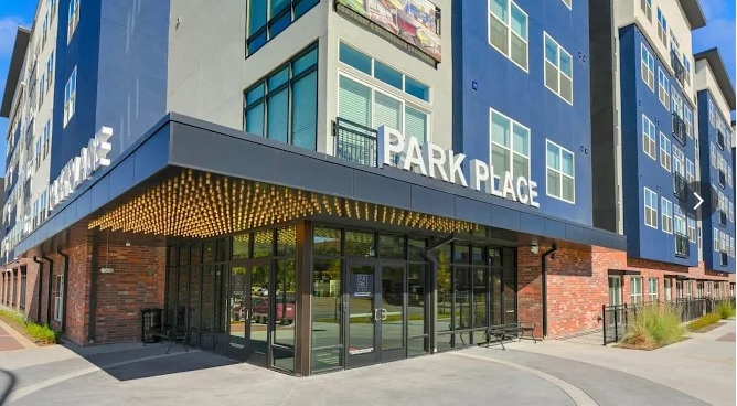 Modern apartment building with blue and gray exterior, large windows, and a sign reading Park Place at Arlington above the glass entryway; landscaped area and sidewalk in front. Experience contemporary living in Arlington.