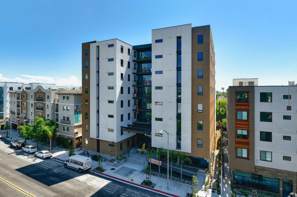 A modern mid-rise apartment building with white and beige facades, large windows, and a central glass entrance, surrounded by palm trees and neighboring residential buildings along a sunny street.