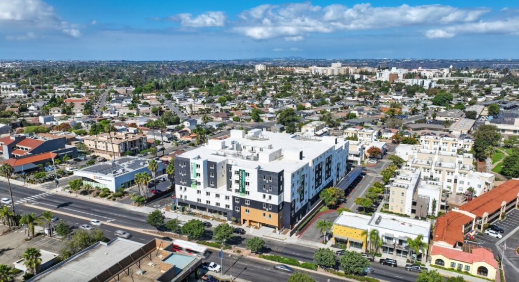 Aerial view of a modern multi-story apartment building surrounded by streets, palm trees, and a mix of residential and commercial buildings, with a cityscape stretching into the distance under a partly cloudy sky.