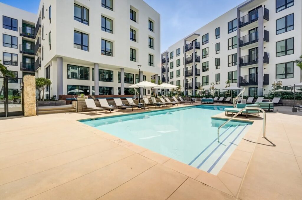 Modern Union Grantville apartment complex courtyard with a rectangular outdoor swimming pool, surrounded by lounge chairs, umbrellas, and tall white buildings featuring large windows and balconies under a clear blue sky.