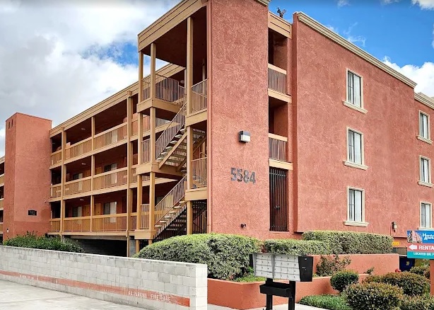 A three-story, red stucco apartment building—Hardy Avenue Apartments—with exterior staircases and balconies stands at 5584. Mailboxes and a low concrete wall are in front, with bushes and a cloudy sky in the background.