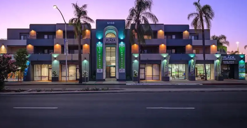 Modern two-story building with glowing Plaza Apartments signage and Dental Office at dusk, surrounded by palm trees and illuminated exterior lights, facing an empty street.