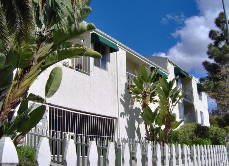 A white apartment building in University Gardens with green awnings and balconies is surrounded by tropical plants and trees, behind a white picket fence, under a sunny blue sky with some clouds.
