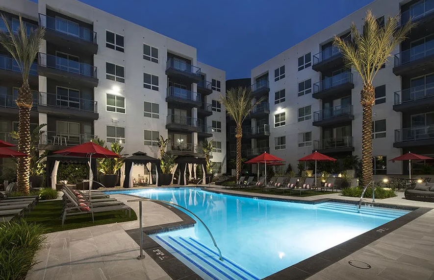 Vora Mission Valley offers a modern apartment complex with multiple stories, large windows, and balconies overlooking a well-lit outdoor swimming pool. Poolside chairs and red umbrellas line the deck, surrounded by palm trees and evening lighting.