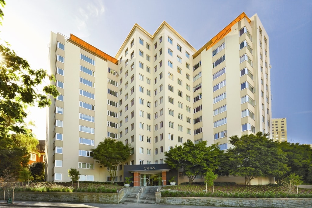 A large, modern apartment building with cream-colored walls, many windows, and a V-shaped design. Trees and shrubs line the entrance, which has steps leading up to a sign that reads Celio Apartments.