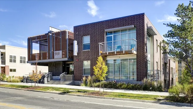 Modern two-story apartment building with large windows, brick and metal exterior, a balcony, and steps leading to the main entrance. The address 910 is displayed by The Gaslamp boulder near the entrance. Trees and a sidewalk are visible in front.