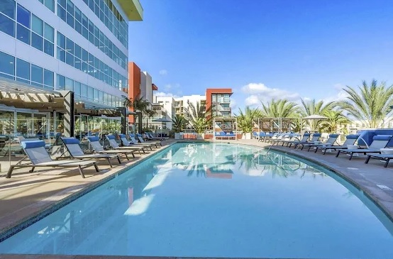 A large outdoor swimming pool surrounded by lounge chairs and palm trees sits beside the modern, multi-story glass building of The Vive Collection San Diego under a bright, clear blue sky.