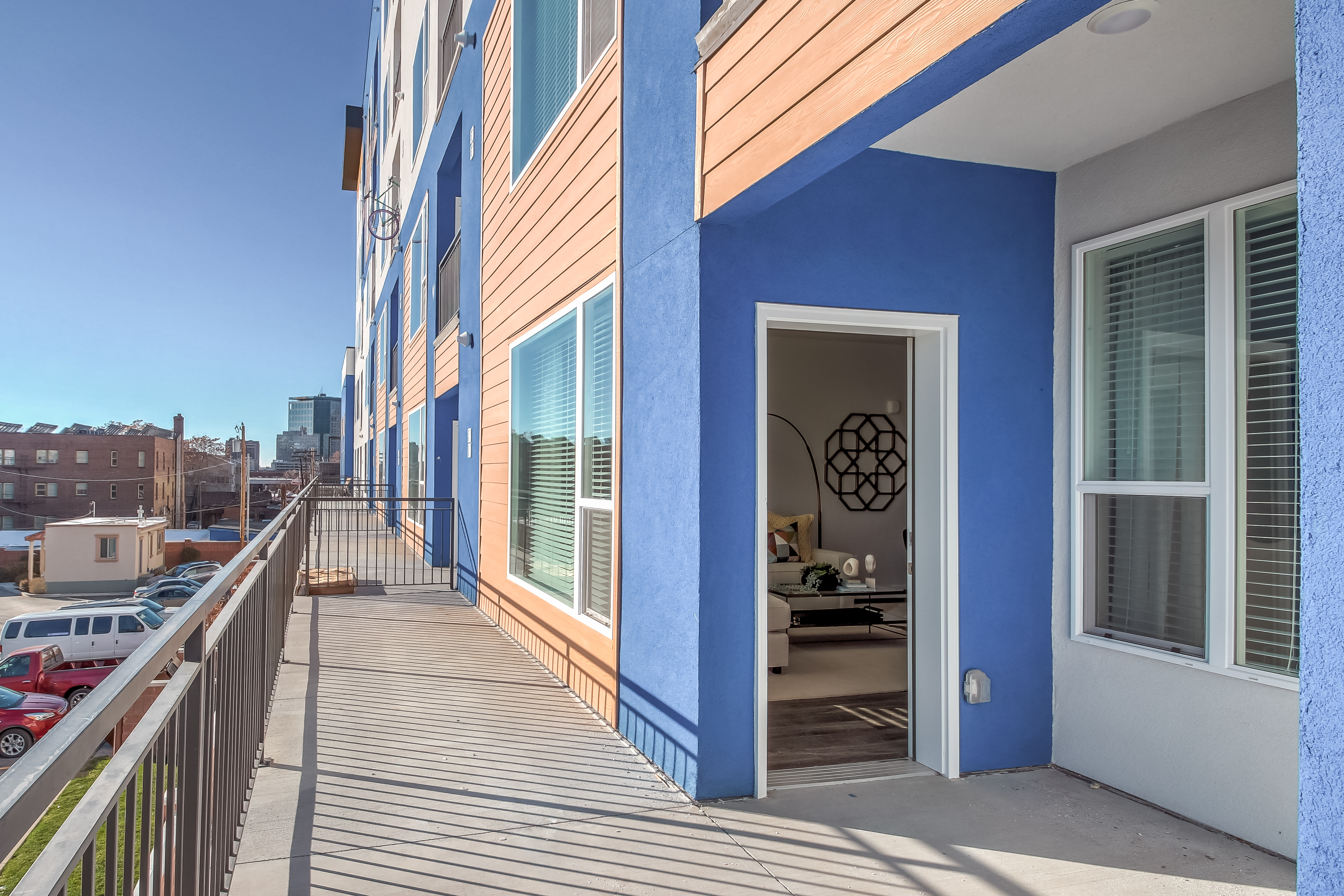 A modern apartment building in Moda Bonneville, Salt Lake City, features a blue and wood-accented exterior, a long balcony with metal railing, and an open door revealing a stylishly furnished living space. Nearby buildings are visible in the background.