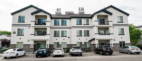A three-story white apartment building with balconies and dark trim at Crestwood Provo, several parked cars in front, viewed from a parking lot on a cloudy day.