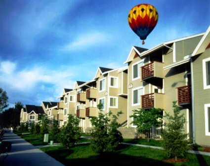 A colorful hot air balloon floats in the blue sky above Morrison Park Apartments Boise, where modern three-story tan buildings with balconies are lined by young trees and a welcoming sidewalk.