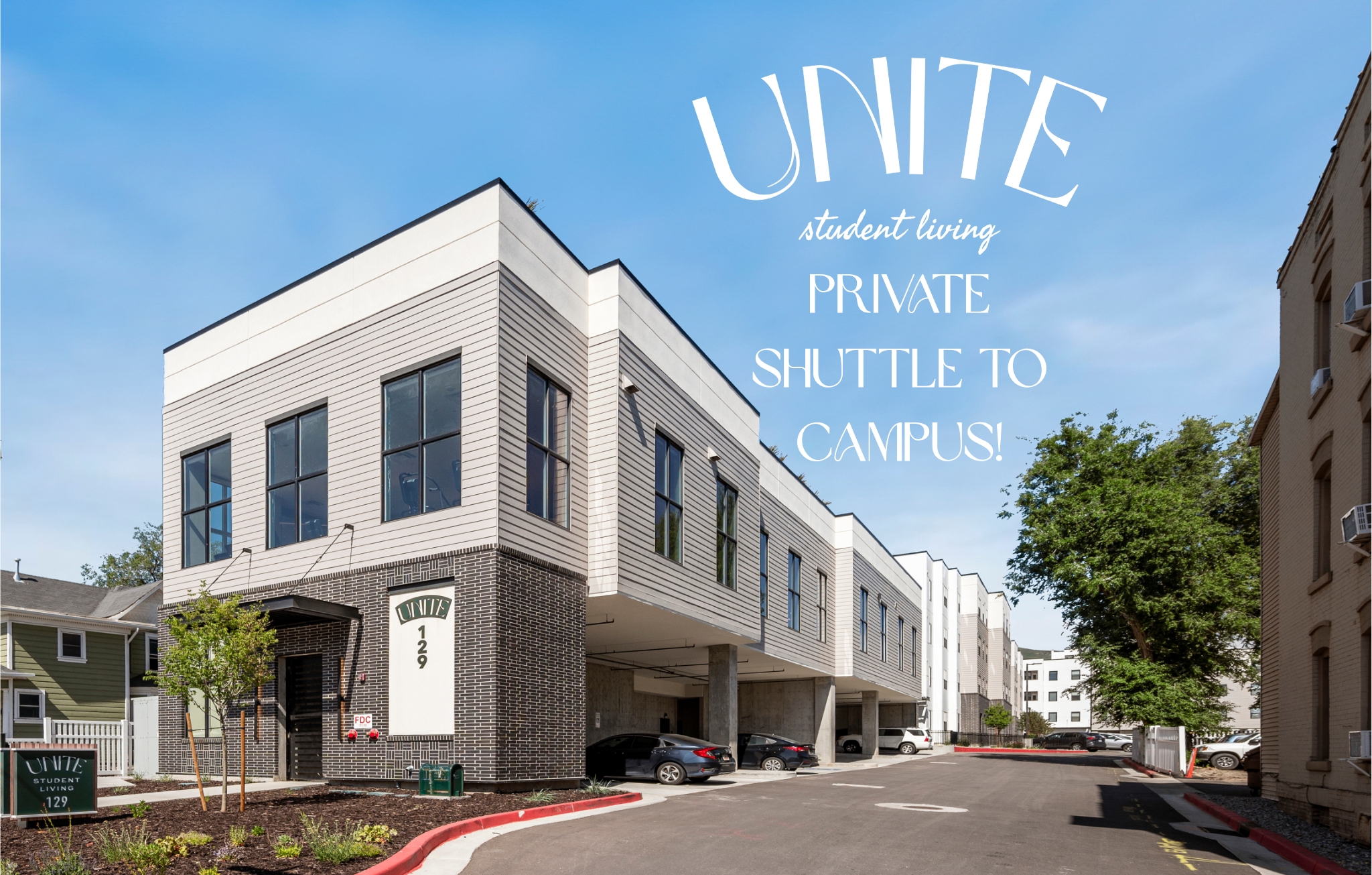 Modern apartment complex with white and gray exterior, labeled UNITE student living and Private shuttle to campus! Several cars are parked under the building at Unite Student Housing Salt Lake City, with trees and clear blue sky in the background.