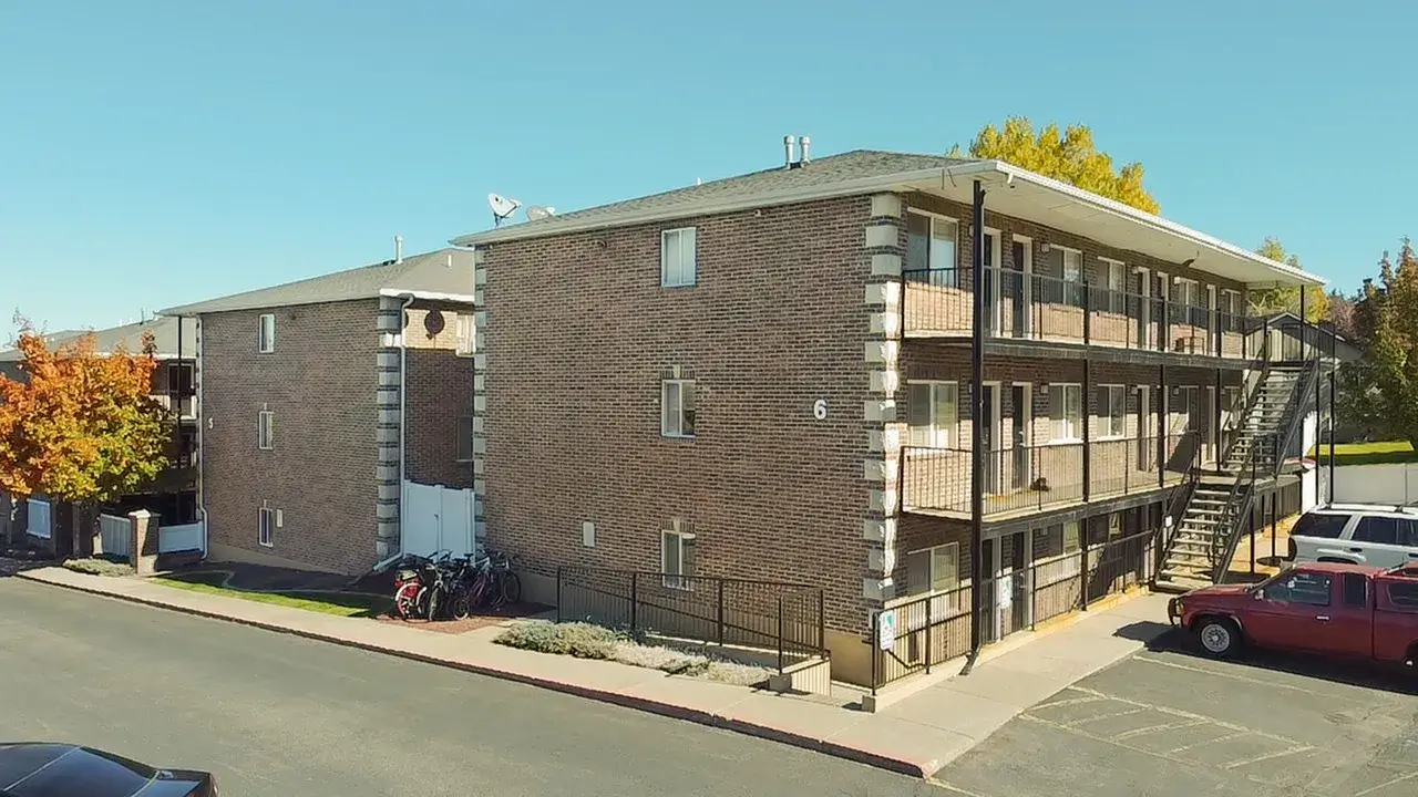 A three-story brick apartment building at University Courtyard Rexburg features exterior staircases and balconies on a sunny day. Several parked cars, bicycles near the entrance, and autumn-colored trees complete the scene.