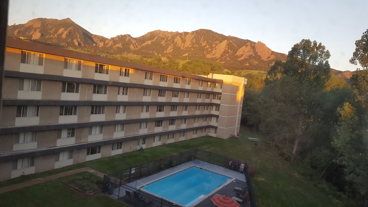 A four-story hotel building, reminiscent of Creekside Apartments Boulder, features a fenced outdoor swimming pool at sunset, framed by tall green trees and sunlit mountains in the background beneath a clear sky.
