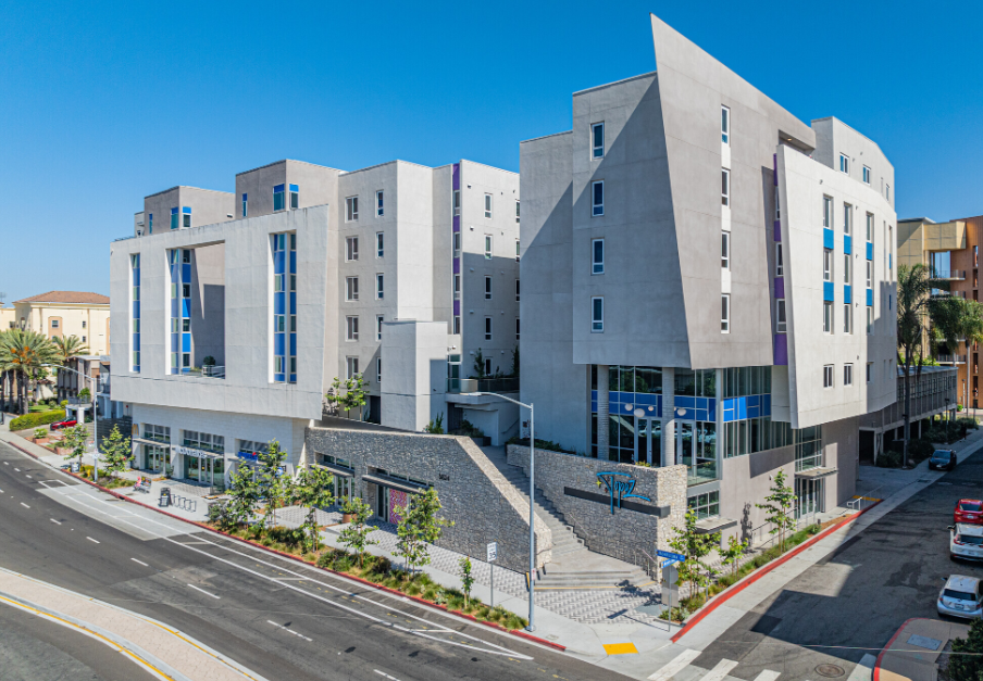 A modern, multi-story apartment building with geometric architecture and large windows sits on a corner in a sunny urban area, surrounded by palm trees, parked cars, and clear blue skies.