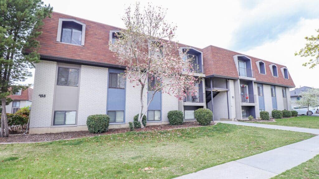 Liberty on Eighth Provo features a three-story apartment building with a brown mansard roof, white and blue-gray exterior walls, several windows, shrubs, a small tree, and a sidewalk in front.