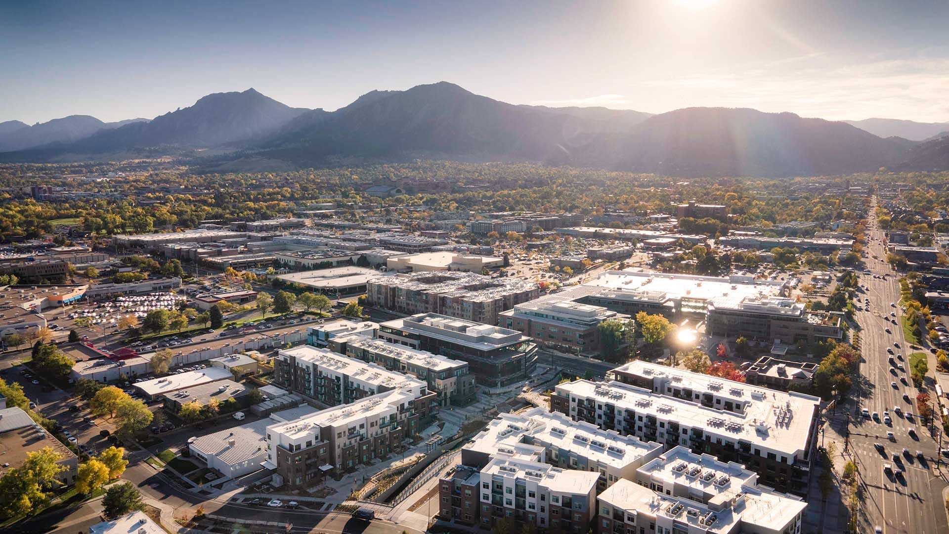 Aerial view of Boulder with modern buildings, tree-lined streets, and mountains in the background under a bright, clear sky with the sun shining—Reve Boulder Apartments offers prime living in this stunning setting.