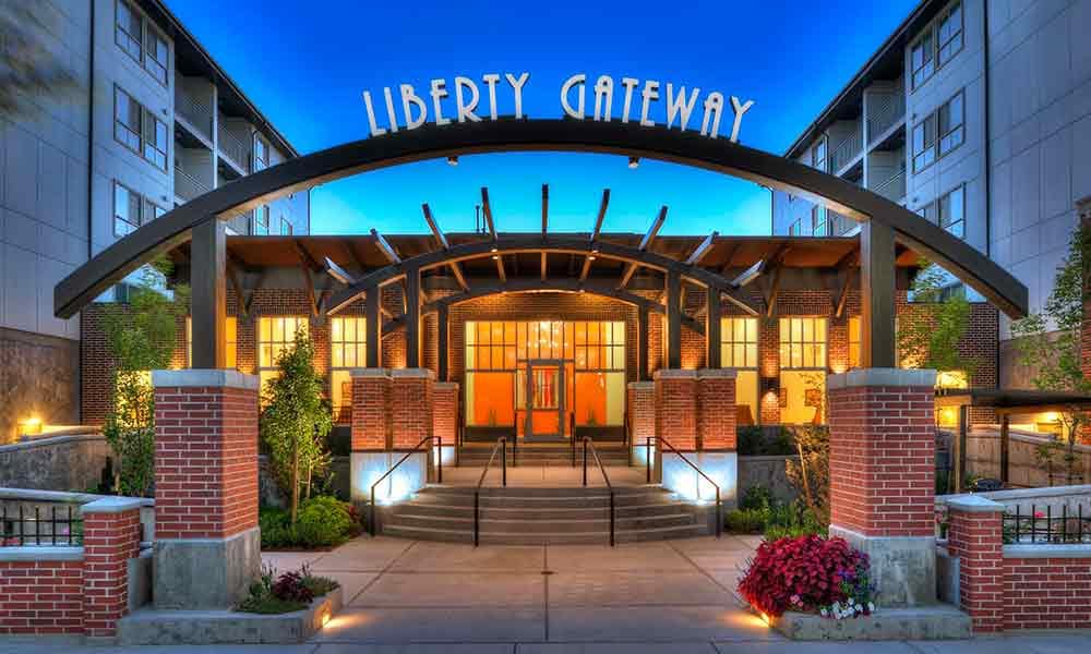Entrance to Liberty Gateway Salt Lake City apartments at dusk, featuring a large metal arch with the name above, brick columns, steps, lush plants, and warm lighting that welcomes visitors.
